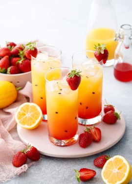 Three glasses with pretty ombre colored Strawberry Passionfruit Lemonade, with black passionfruit seeds speckled throughout and cut lemons and strawberries scattered around.