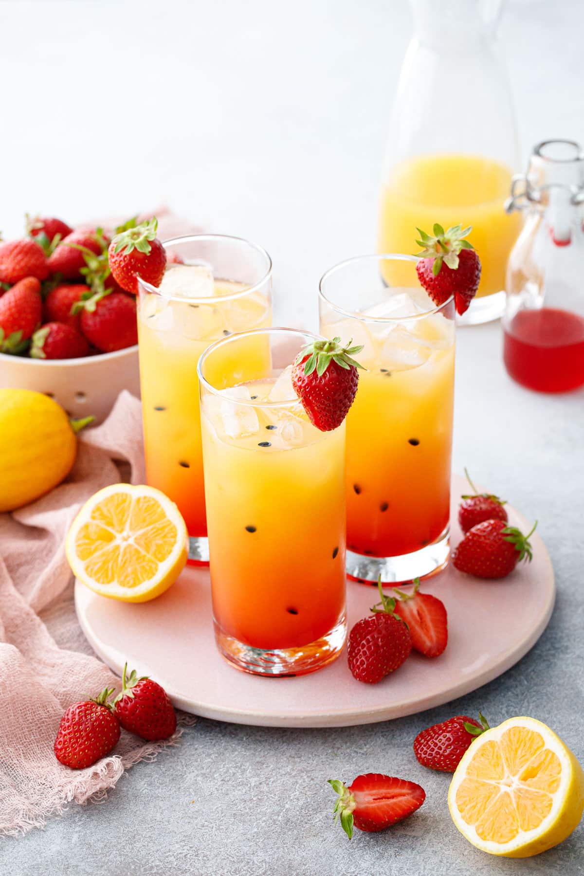 Three glasses with pretty ombre colored Strawberry Passionfruit Lemonade, with black passionfruit seeds speckled throughout and cut lemons and strawberries scattered around.
