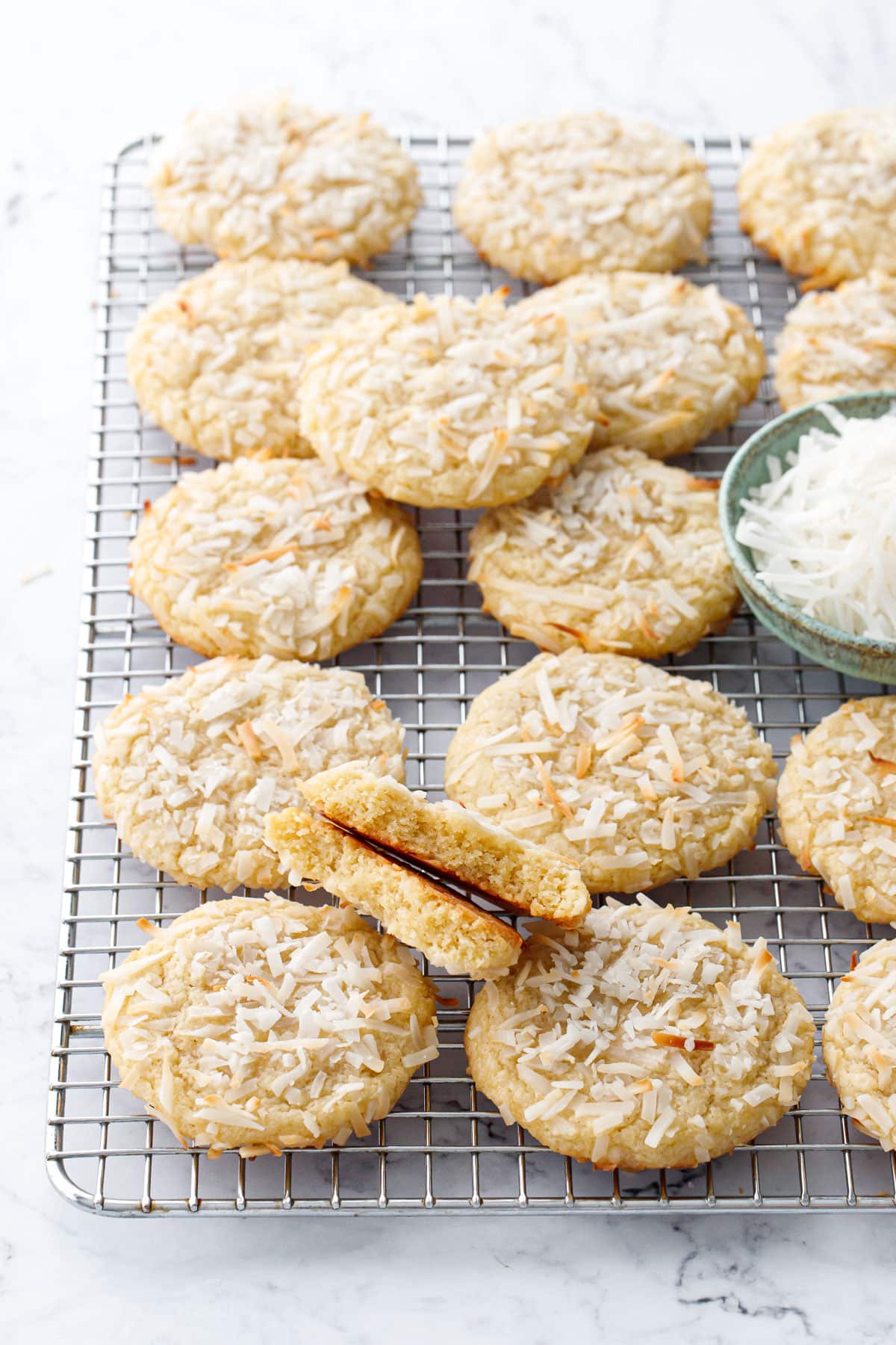 Rows of Toasted Coconut Sugar Cookies on a wire cooling rack with a bowl of coconut, one cookie broken in half sitting on top.