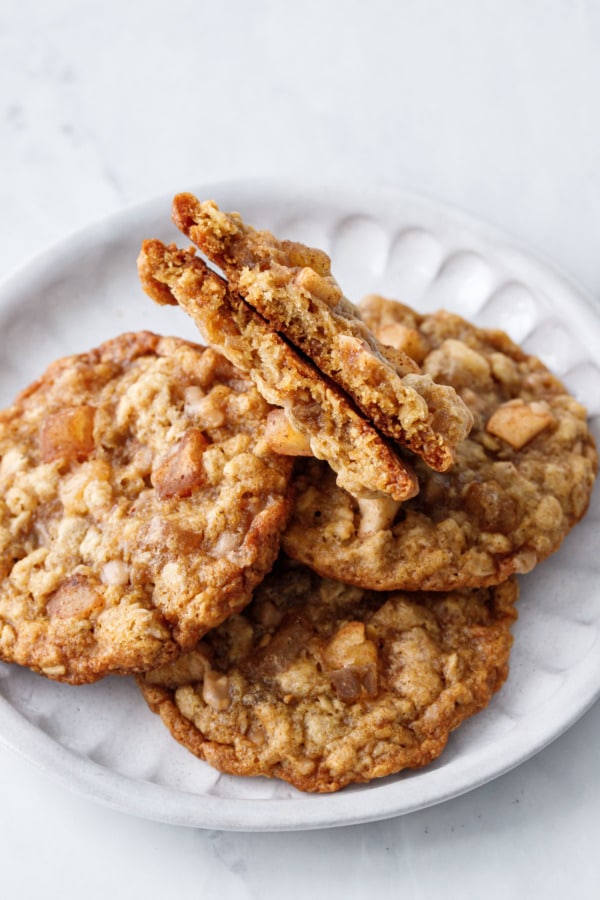 Pile of Toffee Apple Oatmeal Cookies on a white ceramic plate, one cookie broken in half to show the interior texture