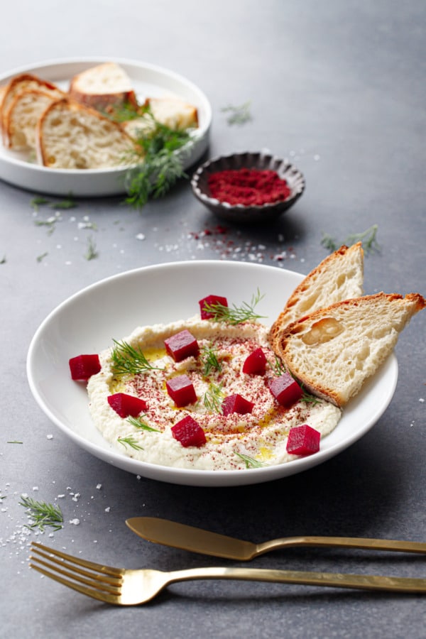 White bowl with whipped almond dip, topped with pickled beets and two slices of fresh sourdough.