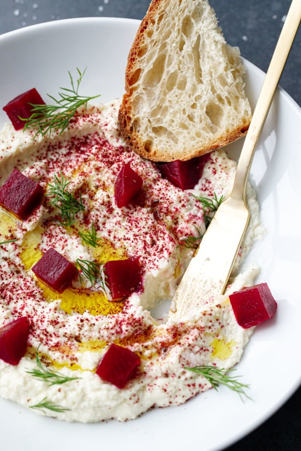 Butter knife in a bowl of whipped almond dip, with olive oil, pickled beets, dill and a slice of sourdough.