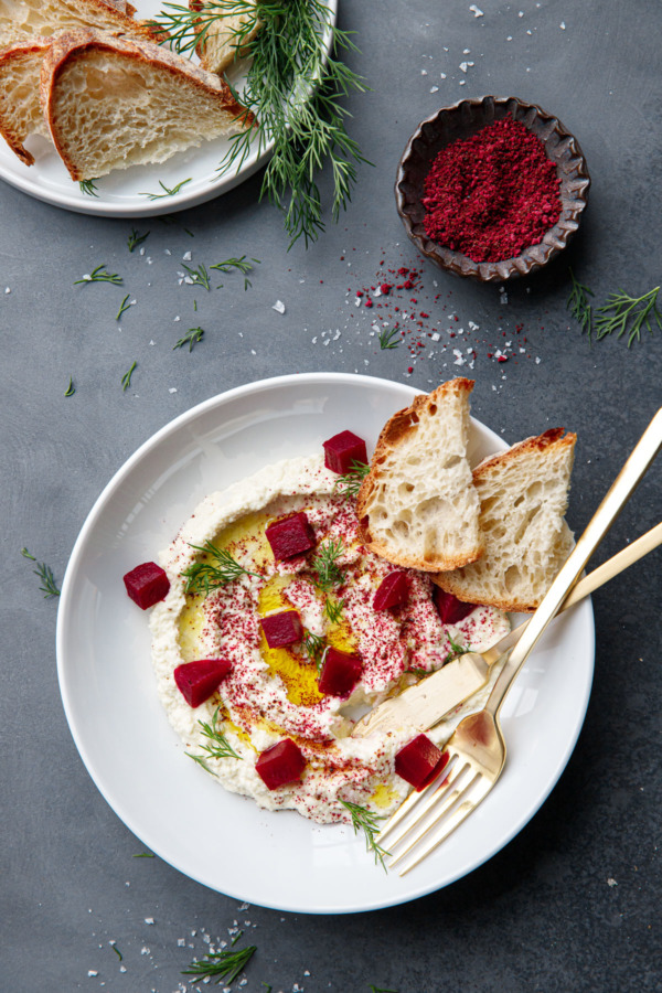 Overhead shot with bowl of whipped almond dip, gold utensils, and bowl of sumac and beet powder and fresh sourdough slices.